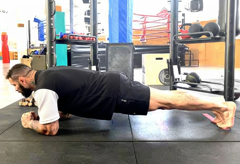 Uomo in posizione di plank su un tappetino da palestra, con attrezzatura sportiva sullo sfondo.